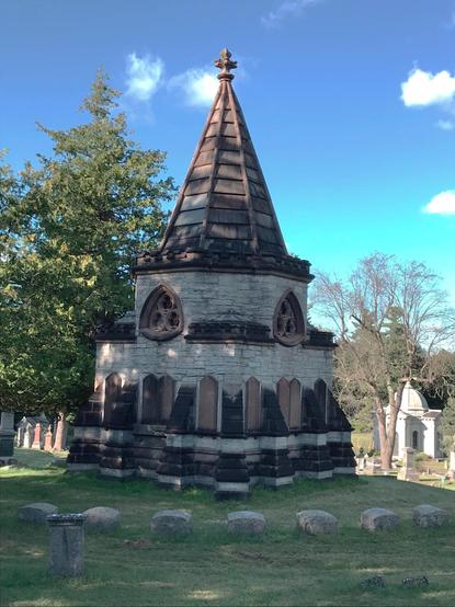 A mausoleum made of blocks of limestone with conical roof, windows, and ornamentation made of brown cast stone. The second level has windows in a trefoil within Reuleaux triangle design. Around the building are gravestones, and another mausoleum can be seen more distantly in the background. The sky above is bright blue with few clouds, but the building is mostly shaded by trees not seen in the image.