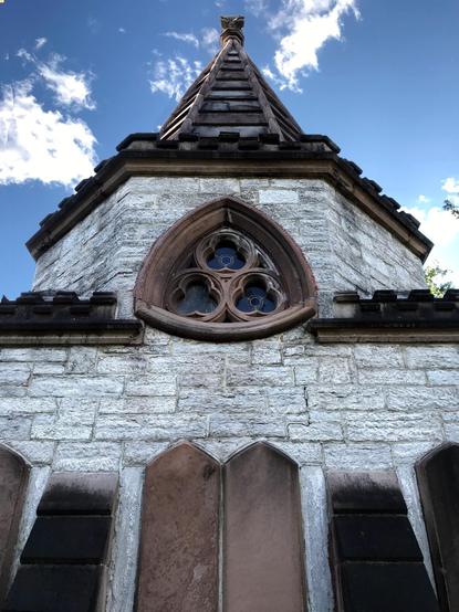 Looking up at the side of a conical-roofed building made of large blocks of limestone with details of brown cast stone. At the center of the picture is a fancy window made of trefoils arranged as a larger trefoil within a Reuleaux triangle. Beyond the building is bright blue sky with a few clouds.