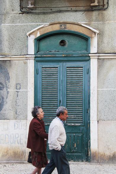 In Olhao, Portugal, an older couple in  simple conservative attire walk past a two-wing wooden door, painted dark turquoise, of an abandoned residential building with gray stone walls. On the left part of a mural can be seen that celebrates Otelo Saraiva de Carvalho, a Portuguese military officer and the chief strategist of the 1974 Carnation Revolution in Lisbon who is still an icon for activists of the left in Portugal. As part of that mural, the word "ADORES" can be seen just to the couple's left. It is however just the ending of the word "trabalhadores" (workers). 

In Olhão, Portugal, geht ein älteres Ehepaar in einfacher, konservativer Kleidung an einer zweiflügeligen, dunkel türkis gestrichenen Holztür in einem verlassenen Wohnhaus mit grauen Steinwänden vorbei. Links ist ein Teil eines Wandgemäldes zu sehen, das Otelo Saraiva de Carvalho, einem portugiesischen Militäroffizier und Hauptstrategen der Nelkenrevolution 1974 in Lissabon, gewidmet ist, der nach wie vor eine Ikone für linke Aktivisten in Portugal ist. Als Teil dieses Wandbildes ist das Wort „ADORES“ (liebt/verehrt) gleich links neben dem Paar zu sehen. Es ist allerdings nur das Ende des Wortes "trabalhadores" (Arbeiter).