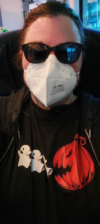 A pale skinned person with auburn hair tied back, wearing sunglasses, a face mask and a black tee with a pumpkin chasing 3 ghosts to eat them on it.