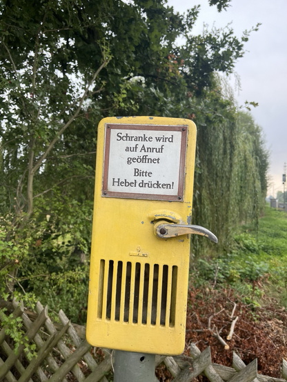 Eine Eiserner Gelber Kasten mit einem Silbernen Hebel an einem Bahnübergang. Aufschrift Schranke wird auf Anruf geöffnet. Bitte Hebel drücken!