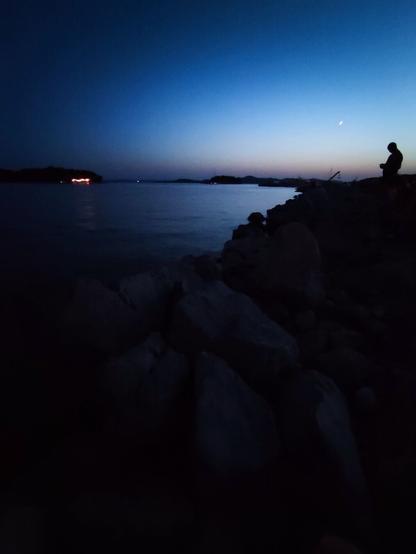 A tranquil evening scene by the water at dusk, with a silhouette of a person standing on a rocky shore. The sky transitions from deep blue to a lighter hue near the horizon. A crescent moon is visible, and distant lights twinkle faintly across the calm water. The mood is peaceful and quiet, with a soft glow of twilight.