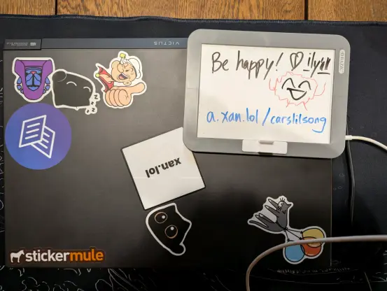 Small whiteboard with drawn text "be happy a.xan.lol/carslilsong" on top of laptop with stickers, like Popeye, cats, and my site xan.lol