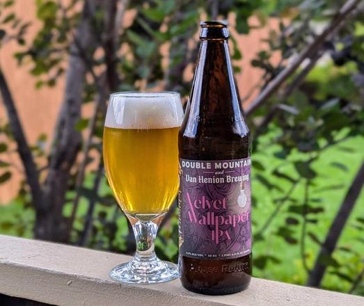 A glass and bottle of Velvet Wallpaper IPA, a collaboration beer from Double Mountain Brewery of Hood River, Oregon, and Van Henion Brewing of Bend, Oregon. The glass and bottle are placed on a porch railing and in the background, blurred out a bit, are shrubbery, a bit of lawn, and fence.