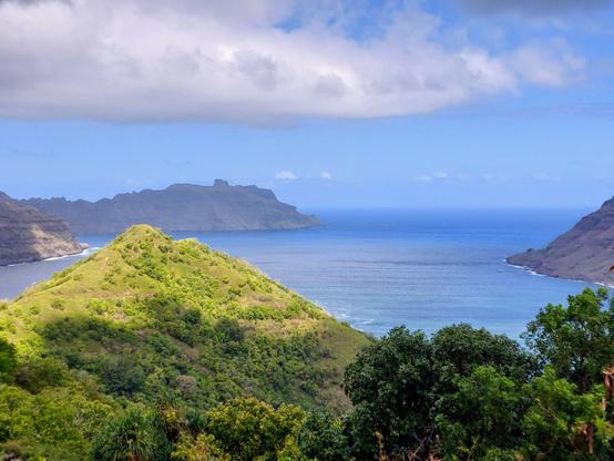 Fotos, die meine Freundin gemacht hat: Im Vordergrund die begrünte  Spitze eines kleinen Berge im Sonnenlicht. Rechts daneben Baumgipfel. Dahinter liegt eine Bucht mit blauem Wasser. Am Himmel ist eine große, weiße Wolke. ~~~ Pictures taken by my friend: In the foreground, the green top of a small mountain in the sunlight. To the right, tree tops. Behind it is a bay with blue water. There is a large white cloud in the sky.