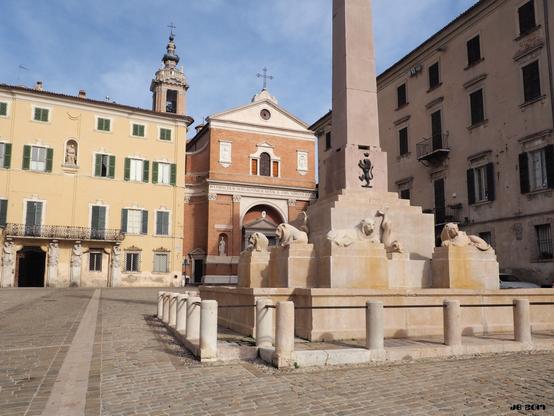 Farbfoto, das die Piazza Federico Secondo in Jesi, Marche, Italien, zeigt. Im Vordergrund ein Brunnen mit Steinskulpturen und Obelisk, im Hintergrund ein stattliches Haus und rechts davon der Dom aus dem 18. Jahrhundert. Der Platz ist dem Stauferkaiser Friedrich II. gewidmet, der in Jesi geboren wurde.