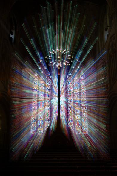 A church window, taken from inside. The light is shinning through the stained glass window. Zooming the lens while taken the photo has given the light an extra appearance of streaming through in lines of light, stopping a short distance in front of the window