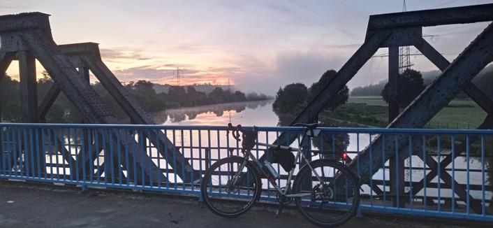 ein Fahrrad steht auf einer Brücke, im Hintergrund geht die Sonne auf. Nebel liegt über dem Fluss
