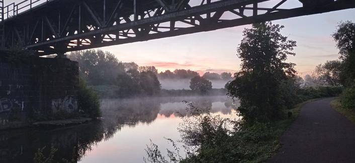 Foto von einem Fluss. Die Wiesen am gegenüberliegenden Ufer sind von Nebel bedeckt.links steht ein Pfeiler im Fluss, der eine Stahl-Fachwerkbrücke trägt, diesen oberen Bildrand rahmt. am rechten Bildrand führt ein Geh- und Radweg am Fluss entlang