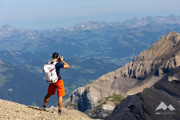 Man from behind, photographing a mountain landscape. He is wearing a dark blue shirt, orange shorts and a white rucksack with dark red details.