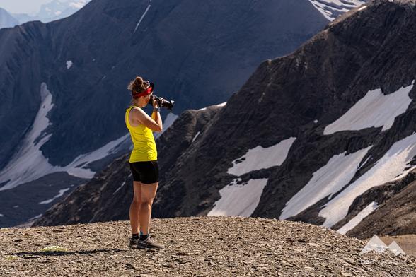 Woman from the side, photographing a mountain landscape with snowfields. She is wearing a red headband, a yellow top, black shorts and is looking straight through her camera.