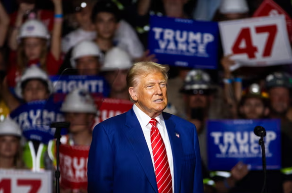 Donald Trump at a political rally wearing a dark blue suit with a bright red tie.

Behind him is a crowd of supporters holding signs. Many of the visible signs say "TRUMP" and there are also signs that say "47".