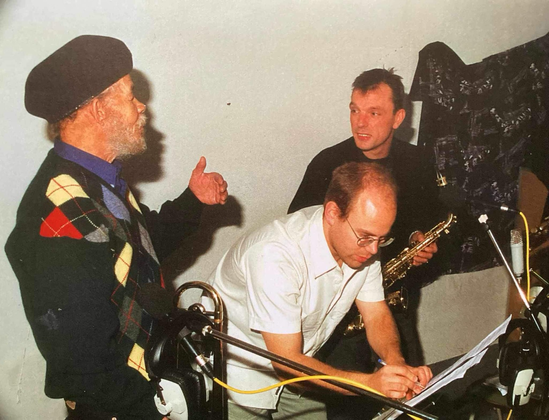 Photo of three persons, holding instruments and discussing something. One of them editing something down on a music stand. Two microphones on stands.