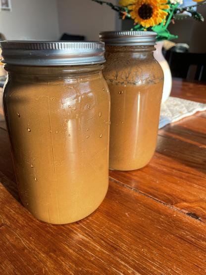 Two mason jars, a wide jar and a narrow jar stand on a wood table. They are filled with water and soil and look like a uniform reddish brown color.