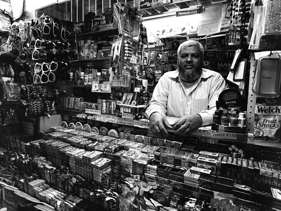 This black-and-white image depicts a man with a beard standing behind the counter of a small convenience store or kiosk. The counter is packed with various products, including candies, gum, snacks, and small everyday items. Behind him, the shelves are filled with a wide range of goods such as sunglasses, batteries, energy drinks, lighters, and other miscellaneous items commonly found in such stores. The man stands with both hands resting on the counter, looking directly at the camera. The space is tightly packed, creating a cozy yet cluttered atmosphere typical of a neighborhood convenience store.