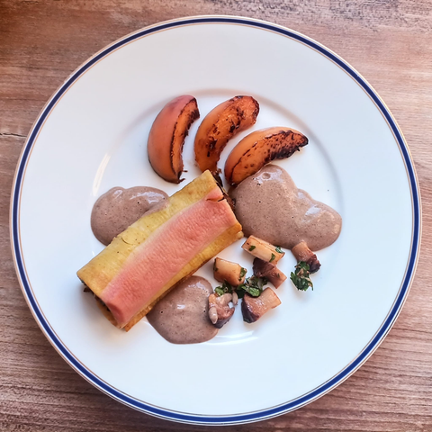 Ein gefüllter Cannoli gelb rot gestreift mit gegrillten Pfirsichstücken, einigen Pilzstücken und Pilzschaum auf dem Teller.