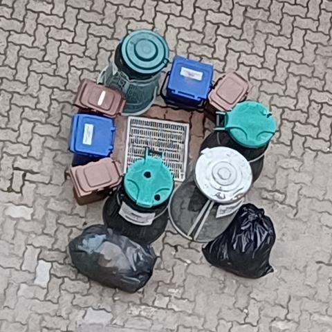 Nine differently shaped and coloured rubbish bins are disposed around a drainer on a paved floor