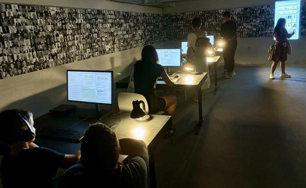 Ein Raum mit mehreren Schreibtischen und Computern, an denen Besucher*innen sitzen und telefonieren. An der Wand im Hintergrund hängen zahlreiche Porträtfotos, die Teil der Installation “Cold Call” sind.