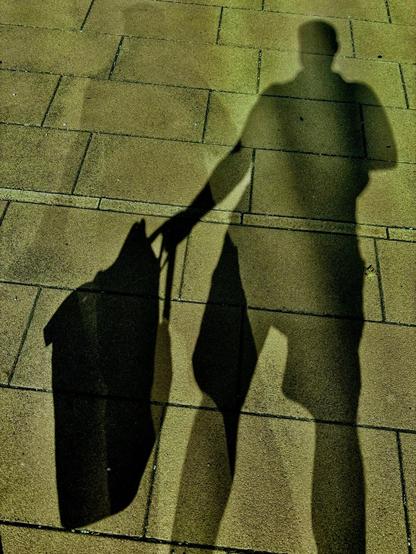 Das Bild zeigt den Schatten einer Person, die auf einem hellen Gehweg aus großen, rechteckigen Pflastersteinen steht. Die Person trägt eine Tasche in der rechten Hand. Der Schatten ist deutlich und lang, durch tief stehendes Sonnenlicht verursacht, und zeigt die Silhouette der Person und der Tasche klar auf dem Boden. Durch eine Lichtspiegelung sieht man den Schatten doppelt.