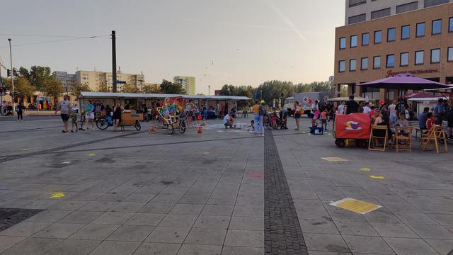 Ein Überblick über die Westseite des Alice Salmon Platzes in Hellersdorf in Richtung der U-Bahn. Es sind einige Stände zu sehen, ein bunter Regenbogen-Fahrradanhänger, ein "Arche Noah" Lastenrad, ein mobiles Fahrradcafé, sowie zahlreiche weitere Stände, die nebeneinander angeordnet sind. Auf dem Boden befinden sich Hinweisschilder, die auf die Bedrohung durch rechte Gruppierungen aufmerksam machen, sowie bunte Händeabdrücke.