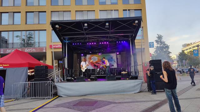 Auf der Bühne befindet sich die Band "The Batteries" aus Hamburg, die Live Rockmusik spielen. Die Bühne befindet sich vor der Sparkasse Hellersdorf, unmittelbar neben dem Bezirksrathaus. Rechts vor der Bühne befinden sich ein Fotograf und ein Metalhead. Ein Securitymitarbeiter ist ebenfalls rechts im Hintergrund zu sehen. Links der Bühne ist ein kleiner abgeschirmter Pavillon, der den Backstage Bereich bildet.