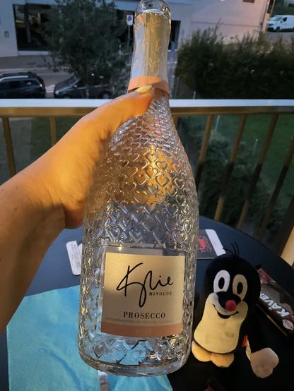 Hand holding an empty bottle of Kylie Minogue Prosecco (glass bottle with small hearts embossed) and Ktreček in the background