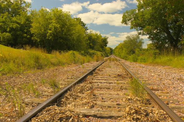 Railroad tracks disappear in the distance. Along side the tracks on both sides are grasses and trees. Blue sky and clouds are visible in the sky.