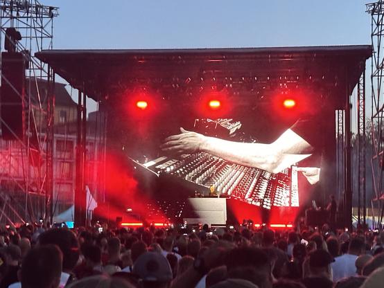 Eine Menge sieht sich eine Live-Musikaufführung mit einem DJ an. Ein großer Bildschirm zeigt eine Nahaufnahme einer Hand, die ein Mischpult manipuliert. Die Szene wird von roten Lichtern gegen einen Dämmerungshimmel beleuchtet.