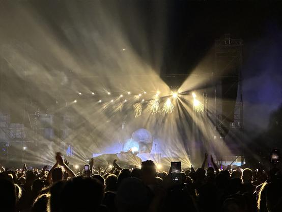 Eine Menge bei einem Konzert mit erhobenen Händen, beleuchtet von dramatischen Bühnenlichtern und einer Kulisse mit einem großen, leuchtenden Objekt.