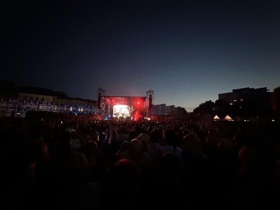 Eine große Menschenmenge versammelt sich bei einem Open-Air-Konzert, mit einer Bühne, die mit roten Lichtern beleuchtet wird, und einer großen Leinwand, die Bilder zeigt. Die Atmosphäre ist lebendig, da die Menschen die Aufführung in der Abenddämmerung genießen, mit Gebäuden im Hintergrund.