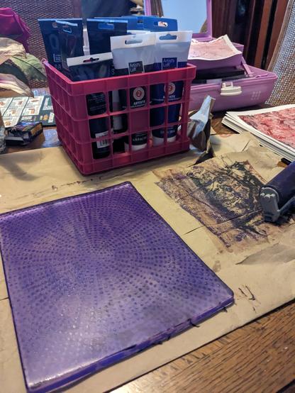 A purple print in progress on a gel plate sitting on top of a paper bag in front a small crate of paints.