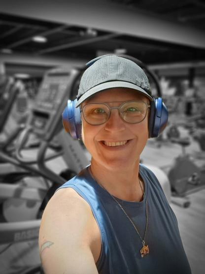 Selfie of a person staving in the gym. Smiling at the camera. Used a filter to have me in color and the background is black and white. Wearing a blue top, black cap, blue headphones and blue-rimmed glasses. In the background are several cardio machines.