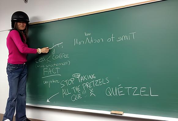 Me, wearing motorcycle helmet, standing on a table & crossing out the word, lies, on chalkboard.