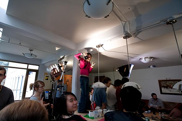 Me, standing on a table, surrounded by filming cast & crew & gear - taking photos with my DSLR in one hand & flashing a peace sign with my other hand.