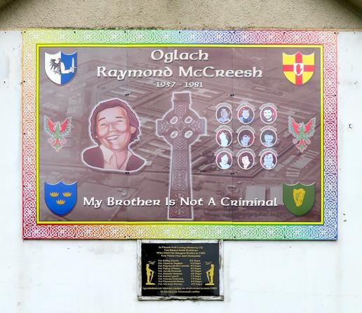 printed board and below it a plaque, on a wall painted white; the board has the crests of the four provinces in the corners, and between the them "Oglach Raymond McCreesh" and "My brother is not a criminal" and two phoenixes; in the centre a larger portrait of one person and nine smaller portraits in a grid are on either side of Celtic cross