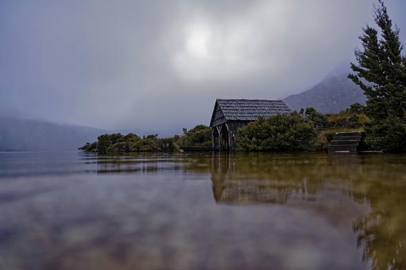 The foreground is the water and can see the pebbles under the water. Centre of the image is the iconic Boatshed of Dove Lake. The background has low clouds obscuring the picturesque cradle mountain.