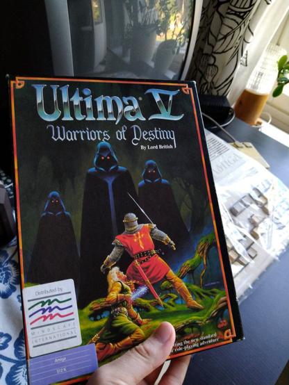 Esko holding the box for Ultima V. The cover depicts hooded figures threatening a chainmail clad hero whose tabard bears a golden anhk. At the foreground, another adventurer has taken a magic arrow to the liver. Amiga 500 is partially visible behind the box.