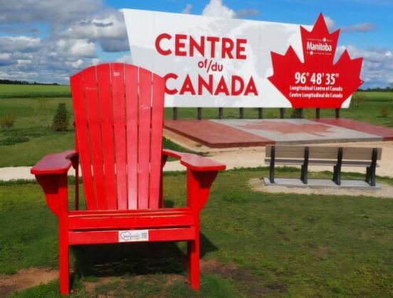 A large roadside sign marks the longitudinal center of Canada on the Trans Canada Highway near Tache, Manitoba,  in front of the Sign is a large Canadian flag on the ground, and an oversized Adirondack chair painted bright red.