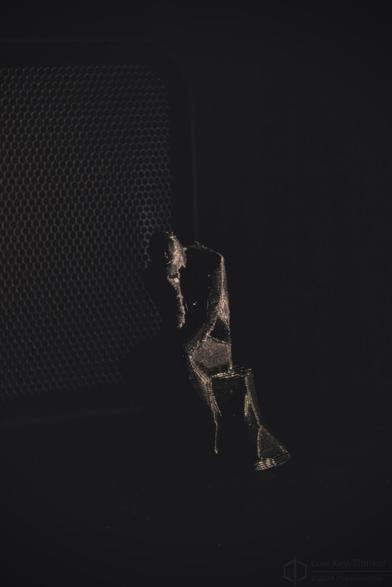 Low key close up photo of a 3D printed low polygon thinker statue where the layer lines are catching light. A speaker grill in the background