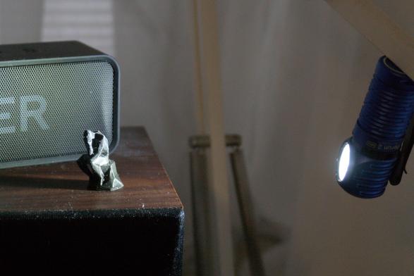 BTS of the first picture. The 3D printed figurine is shot on a faux wood speaker, a bluetooth speaker behind it and a small flashlight providing lighting hanging from a desk lamp frame.