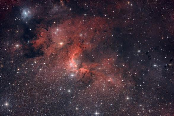 At the center of a dense star field, the cave nebula is a cloud of red ionized hydrogen gas forming a C shape with the opening oriented towards the top-right of the image.

A large dark cloud of interstellar dust located on the left of the nebula blocks the light from the emission nebula and background stars. Multiple smaller dark clouds can be seen around the nebula.

A large orange star is located between the center of the nebula and the large dust cloud.

In the top left corner, the light from two large stars reflect on the surrounding interstellar dust, forming a light blue halo.
