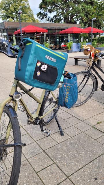 Ein grünes Fahrrad mit blauer Lenker- und Gepäckträgertasche. Das Fahrrad ist von vorne zu sehen. Dahinter ein schwarzes Fahrrad von dem nur der vordere teil zu sehen ist. Im Hintergrund rote Sonnenschirme vor einem Café und sommergrüne Laubbäume.