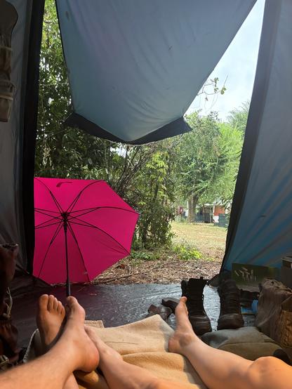 Ein Zelt von innen. Der Eingang ist als Vordach hochgebunden. Im Eingang ein pinker Regenschirm. Innen zwei Paar nackte Beine.