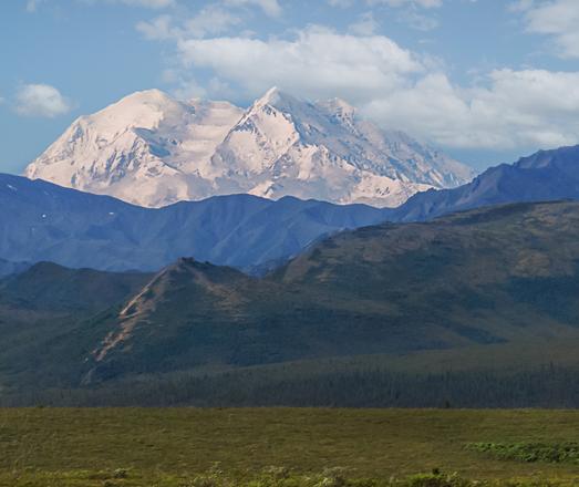 Photo of Denali from 70 miles away. Snow covered mountain in the background. lower mountains and hills in the middle and a grassy plain in the foreground.