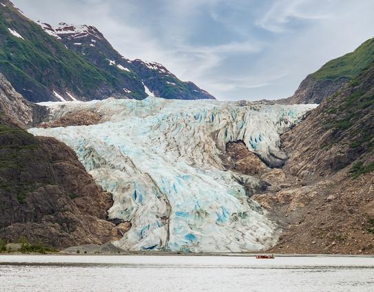 The foot of a glacier sloping down to the water. Mountains rising from the sides of the glacier. Wispy clouds in the sky.