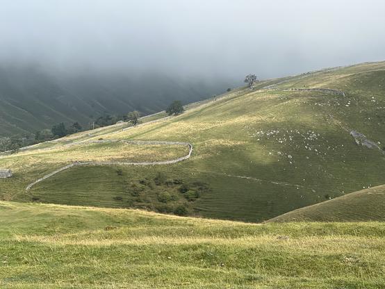 A scenic landscape featuring rolling green hills, dry-stone walls, and patches of fog above. The terrain is gently undulating, with a few trees scattered throughout.