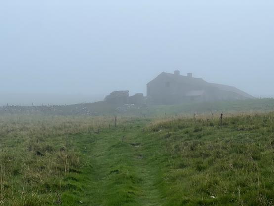 A foggy landscape featuring a grassy path leading towards an old stone building partially obscured by mist, with additional ruins visible in the background.