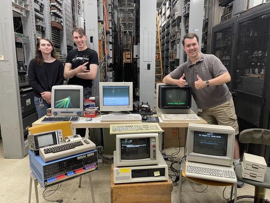 3 people smiling proudly and standing behind/leaning on some older computer models out on display, all turned on with something on the screen. there's aisles of hardware behind them stretching on seemingly forever