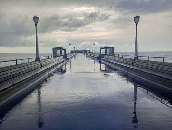 Looking down a wet, concrete, pier with grey sea and stormy clouds