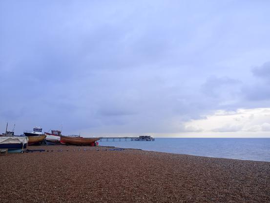 Looking over a shingle beach to a grey sea, on horizon a pier, on left of beach are fishing boats. Very dark grey clouds with a hint of brightness on horizon